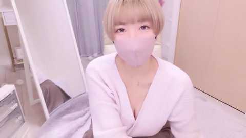 Media: Video of an Asian woman with short blonde hair, wearing a white bathrobe and a pink face mask, seated on a white chair in a modern bathroom with light-colored walls and a white sink.