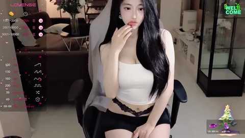 Media: Video of an Asian woman with long black hair, wearing a white crop top and black lace panties, sitting on a black chair in a modern, brightly lit living room.