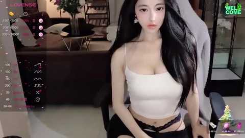 Media: Video of a young East Asian woman with long black hair, fair skin, and a slim physique, wearing a white crop top and black shorts, sitting on a chair in a modern living room with a TV screen displaying a virtual avatar.