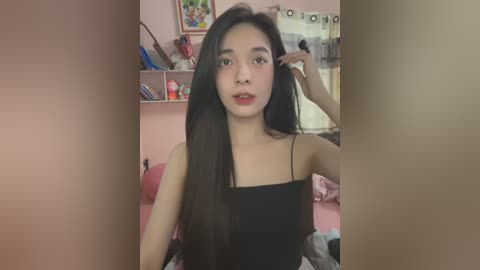 Media: A video of a young East Asian woman with long black hair, wearing a black spaghetti-strap top, posing in a bedroom with pink walls and a framed painting.