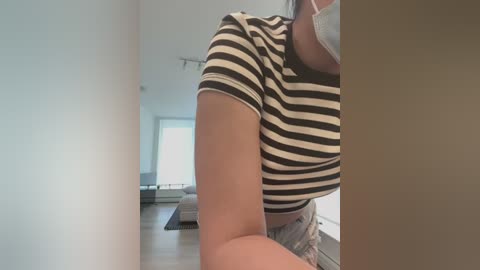 Media: Video of a person with fair skin wearing a black and white striped top, standing in a modern, minimalist living room with light wooden floors and a sofa.