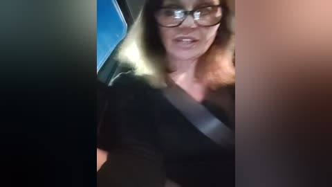 Media: Video of a middle-aged woman with glasses, shoulder-length brown hair, wearing a black top, driving a car with a seatbelt. Background shows blurred car interior and street light.