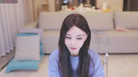 Media: Video of a young Asian woman with long black hair, light skin, and fair complexion, wearing a light blue top, sitting on a transparent chair in a modern, minimalist living room with beige couch and blue cushion.