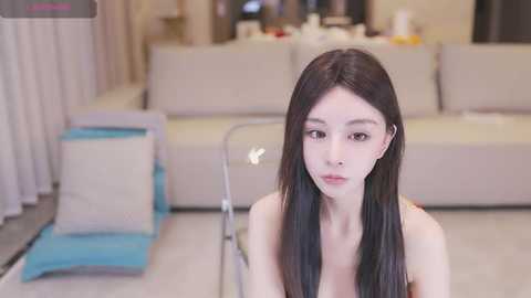 Media: Video of a young East Asian woman with long black hair, pale skin, and small breasts, sitting in a modern living room with beige couches, a blue chair, and a white wall.