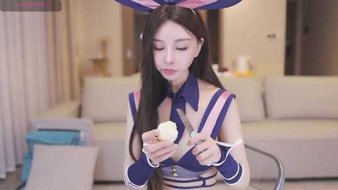 Media: Video of an Asian woman with long black hair in a bunny costume, holding a knife, in a modern living room with beige couches and neutral decor.