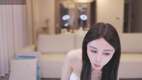 Media: Video of a young East Asian woman with long black hair and fair skin, wearing a strapless white dress, sitting on a beige sofa in a modern, softly lit living room with cream curtains and a mirror.