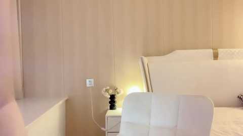 Media: Video of a minimalist, beige-walled bedroom featuring a modern, white upholstered bed, a small white nightstand with a lamp and flowers, and a sleek, white chair.