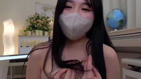 Media: Video of a young East Asian woman with long black hair, wearing a surgical mask and a light gray tank top, in a modern, well-lit room with a vase of flowers, a globe, and a lamp in the background.