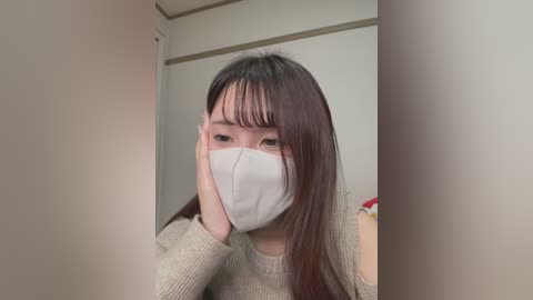 Media: A video of an East Asian woman with straight, dark hair and bangs, wearing a white face mask, seated in a room with beige walls and a red object in the background.