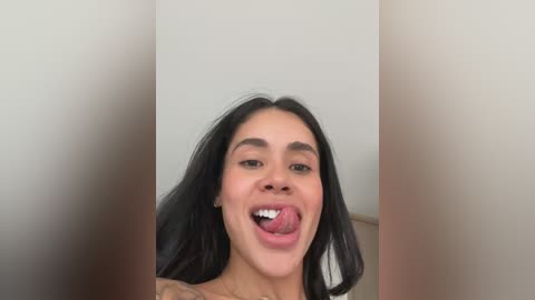 Media: Video of a young woman with long black hair, fair skin, and light makeup, sticking her tongue out. She wears a tattoo on her upper arm. Background is a plain, off-white wall.