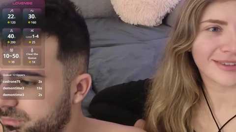 Media: A video of a young, smiling woman with light skin and long, wavy blonde hair, wearing a black necklace, sitting next to a man with short dark hair and a beard, both on a gray bed with a pink pillow.