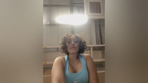 Media: Video of a young woman with curly hair, wearing a light blue tank top, standing in a brightly lit, industrial workshop with shelving units and tools in the background.