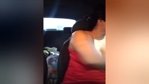 Media: A video showing a person with dark hair wearing a red tank top, seated in the backseat of a car, with a blurred background of a white light and a partially visible bag.