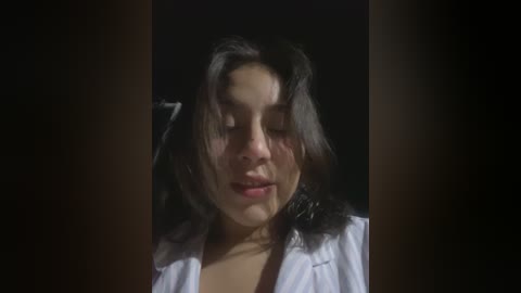 Media: A close-up video of a young woman with long, dark hair and fair skin, eyes closed, wearing a white shirt, set against a dark background.