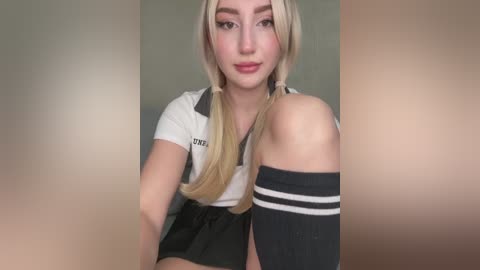 Media: Video of a young woman with fair skin, blonde pigtails, and a white polo shirt with \"XXX\" on the collar, wearing a black skirt and thigh-high black socks with white stripes, in a neutral background.