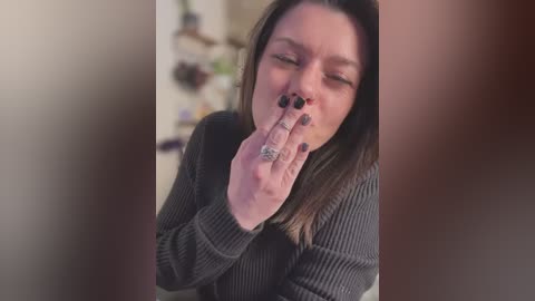 Media: Video of a Caucasian woman with long, straight, brown hair, wearing dark, long-sleeved clothing, holding a finger to her nose, with black nail polish and multiple rings. Background shows blurred kitchen items.