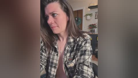 Media: A video of a middle-aged woman with shoulder-length brown hair, wearing a plaid shirt over a pink top, looking thoughtful, in a cozy, cluttered living room with green plants and wooden furniture.