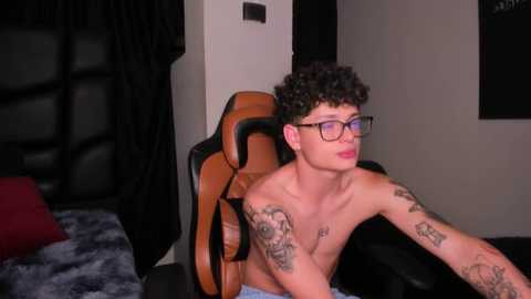 Video of a shirtless young man with curly hair, wearing glasses, sitting in a gaming chair, tattoos on his arms, in a dimly lit bedroom with a bed and black curtains.