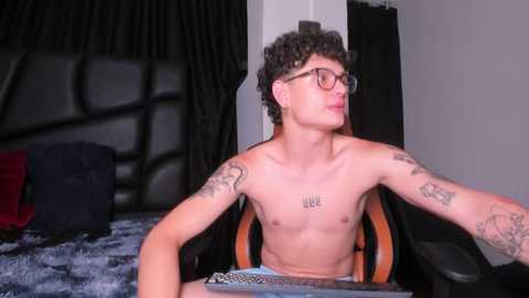 Media: Video of a shirtless young man with curly hair, glasses, tattoos, and a black leather headboard, sitting on a gaming chair in a modern bedroom.