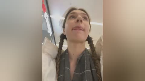 Media: Video of a young woman with light skin and brown hair styled in pigtails, wearing a gray plaid scarf and white shirt, looking upward with a neutral expression. Background shows blurred interior details.