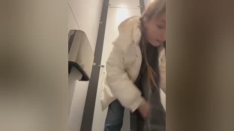 Media: Video of a woman in a public restroom, wearing a white puffy coat and blue jeans, bending over to pick up a tissue, with a gray towel in the foreground.