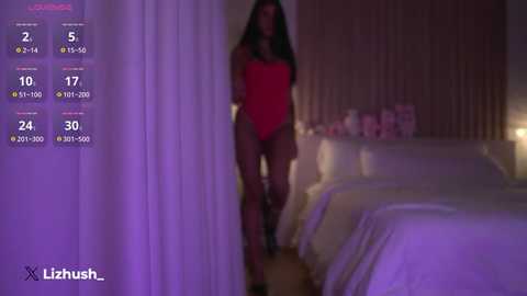 Video of a woman in a red bodysuit, standing in a dimly lit bedroom with a bed and curtains.