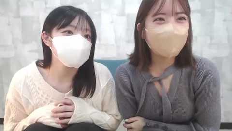 Media: Video of two East Asian women in face masks, one in a white knit sweater, the other in a gray sweater, sitting side-by-side against a white, patterned backdrop.