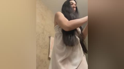 Media: A video shows a woman with long, straight black hair, wearing a white, sheer nightgown, adjusting her hair in a bathroom with beige stone walls and a pink towel hanging on a rack.