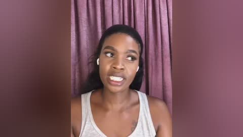 Media: Video of a dark-skinned woman with long black hair, wearing white headphones, a grey tank top, and a light pink wall with maroon curtains in the background.