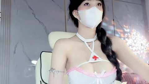 Media: Video of an Asian woman with long black hair in braids, wearing a white nurse costume with a red cross emblem, a face mask, and a silver bracelet, seated in a modern office chair against marble walls.