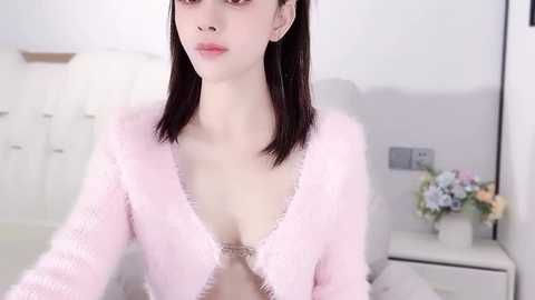 Media: Video of a fair-skinned, slim Asian woman with straight black hair, wearing a pink, fuzzy robe, standing in a minimalist, white room with a flower vase on a white nightstand.