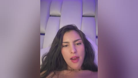 Media: A video of a topless, light-skinned woman with long dark hair and a nose ring, in a white-tiled room, with purple lighting.