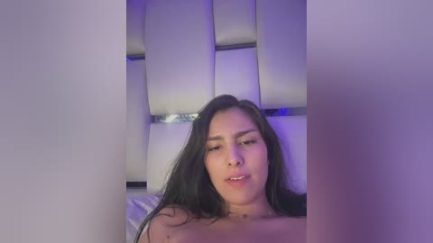 Media: A video of a topless Latina woman with long, dark hair, medium skin tone, and full lips. She lies on a white, padded headboard with a purple light casting a soft glow.