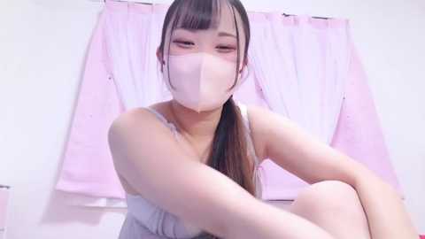 Media: A video of an Asian woman with light skin, wearing a pink mask and white top, sitting on a bed with pastel pink curtains in the background.