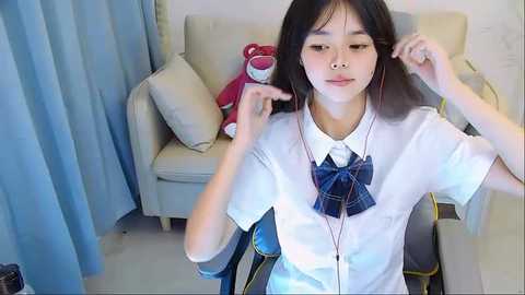 Media: Video of an Asian girl with straight black hair, wearing a white blouse and blue plaid bowtie, adjusting her earphones. Background includes a beige couch, red plush bear, and light blue curtains.