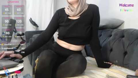 Media: Video of a woman in black leggings and a black crop top with a beige hijab, sitting on a gray couch, with a fitness app overlay on the screen.