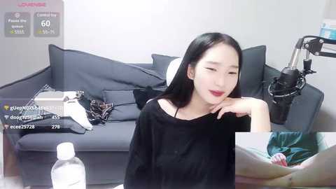 Media: A video of a young East Asian woman with long black hair, wearing a black top, sitting on a gray sofa. Background includes a bottle of hand sanitizer, clothes, and a microphone.