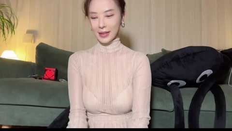 Media: A video of an Asian woman with light skin, brown hair tied back, wearing a beige ribbed turtleneck sweater, sitting on a green sofa in a cozy room with beige walls and a lamp.