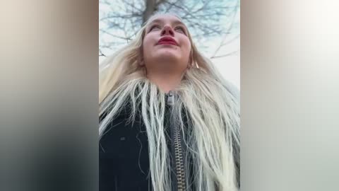 Video of a blonde woman with long hair, wearing a black jacket with a fur trim, looking upwards. Background features bare trees and a cloudy sky.