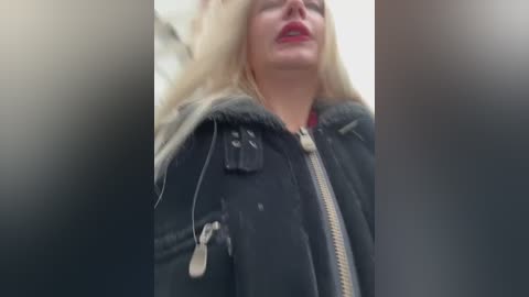 A video of a blonde woman in a black jacket with a fur-lined hood, wearing red lipstick, taken from a low angle, with blurred background.