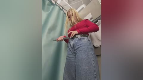 Video of a blonde woman in a red top and high-waisted blue jeans, adjusting a belt in a bathroom with teal and pink tiled walls.