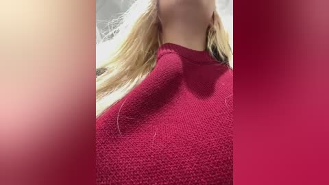 Video of a woman's neck and shoulders, wearing a red knit sweater. Her blonde hair is partially visible. Background features a gradient from beige to pink.