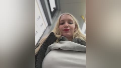 A video of a blonde woman with fair skin and red lipstick, wearing a gray sweater, sitting in an airplane's aisle. The background shows the cabin's white walls and a blurred passenger.