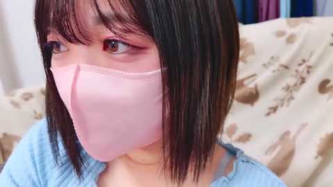 Media: Video of an Asian woman with straight black hair, wearing a pink mask and light blue top, looking up. Background shows a bed with a beige sheet and blue pillowcases.