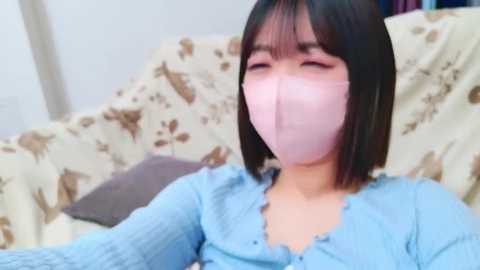 Media: Video of an East Asian woman with short black hair, wearing a light blue ribbed sweater and a pink surgical mask, seated on a beige couch with brown patterned fabric.