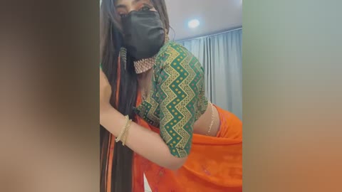 Media: Video of a woman with medium-brown skin, wearing a green and yellow patterned blouse and orange saree, black mask, and black gloves, in a modern indoor setting.