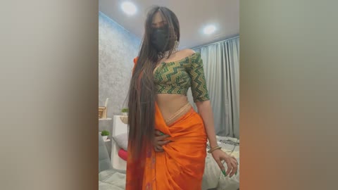 Media: Video of a woman with long, straight black hair, wearing a green off-shoulder crop top, orange sari, and mask, standing in a modern, softly lit bedroom with white walls and a bed in the background.