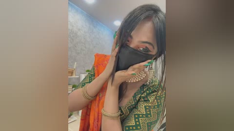 Media: A video of a South Asian woman with medium skin tone, dark hair, and green eyes, wearing a green and gold traditional sari, black face mask, and gold bangles, taking a selfie indoors with a blurred background.