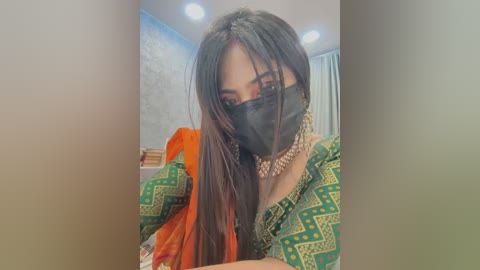 Media: Video of a woman with long black hair, wearing a green patterned dupatta, a black face mask, and a gold necklace. Background shows a blue and white wall, a bookshelf, and a window.