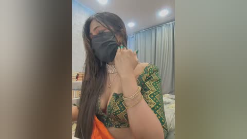 Media: Video of a woman with long dark hair, wearing a green saree with gold designs, a black face mask, and multiple gold necklaces, in a dimly lit bedroom with blue curtains.